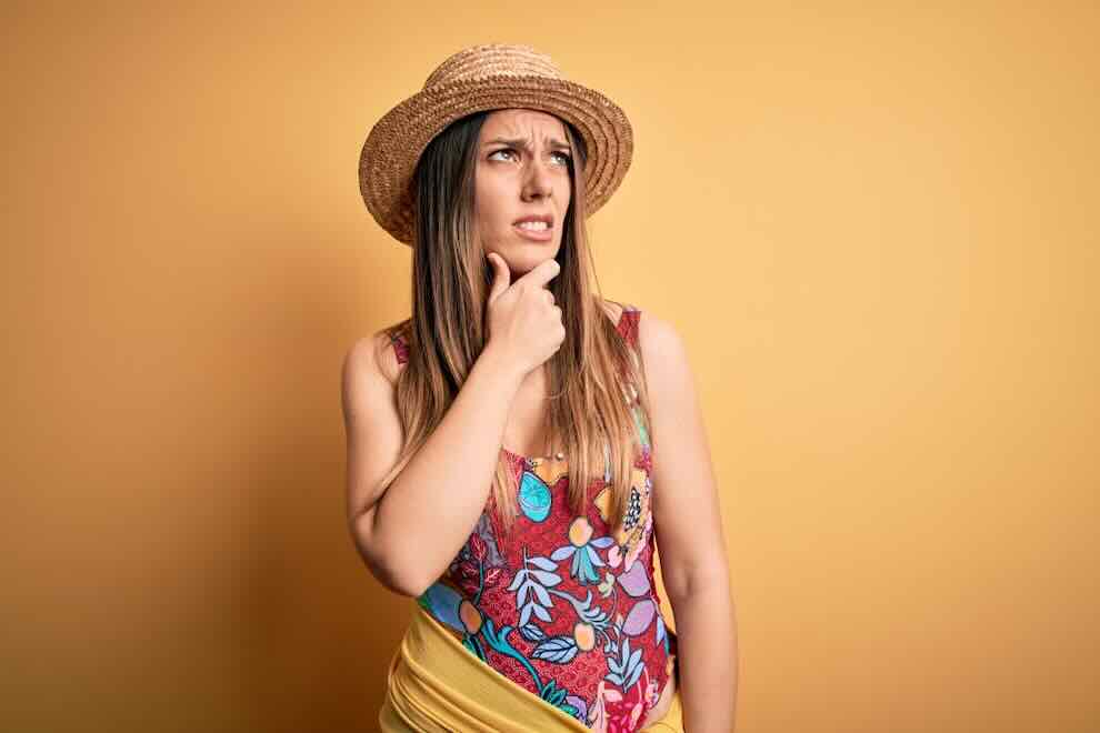 Young blonde woman wearing swimsuit and summer hat over yellow background Thinking worried about a question, concerned and nervous with hand on chin