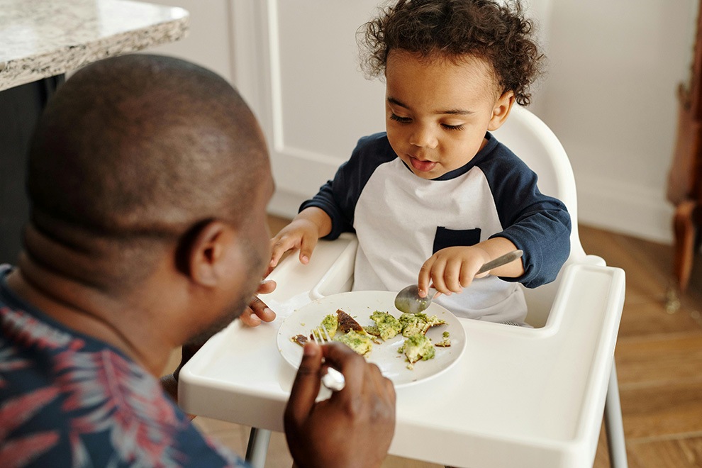 father helping young child in highchair eat healthy food